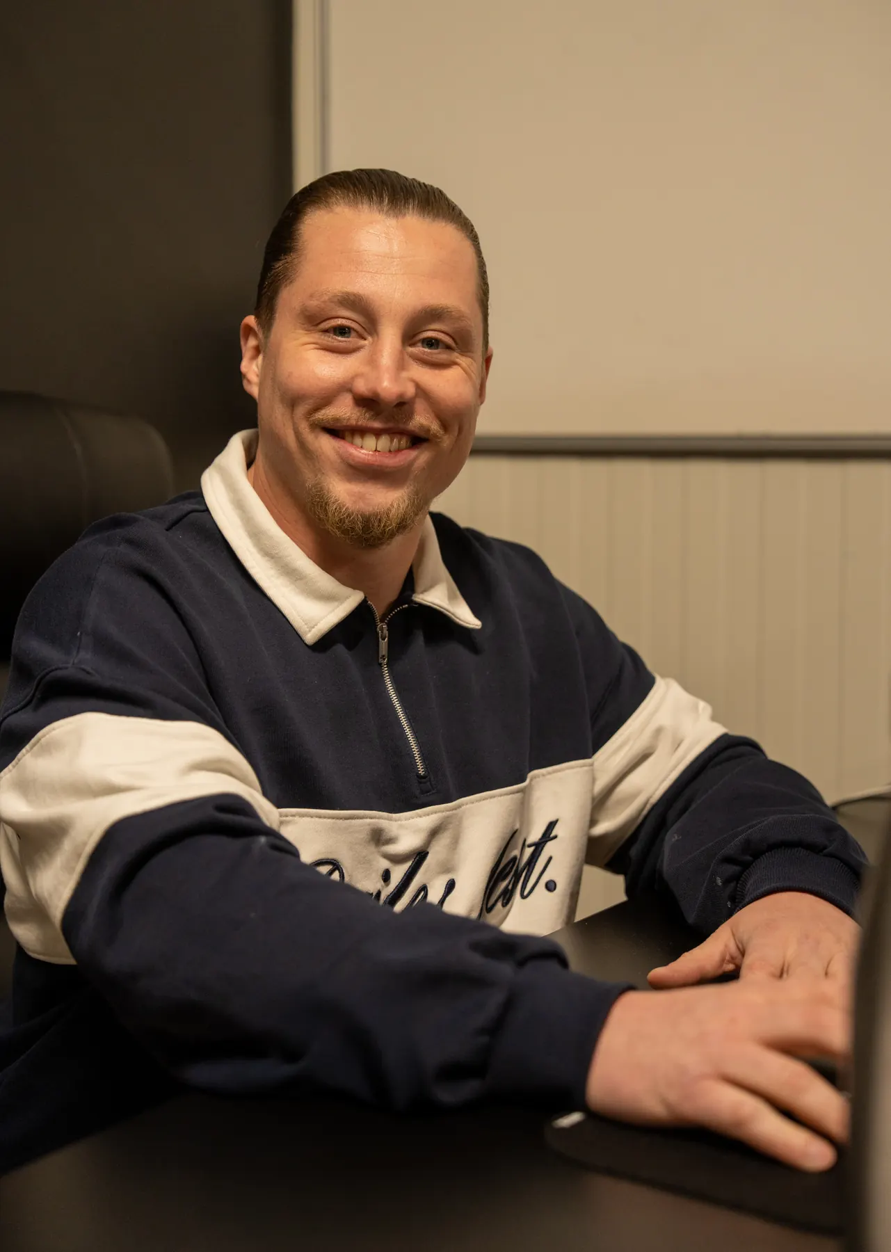 Stevie, eigenaar van VerhuisMeesters, zit lachend aan een tafel in het kantoor, in een donkerblauwe trui met witte kraag