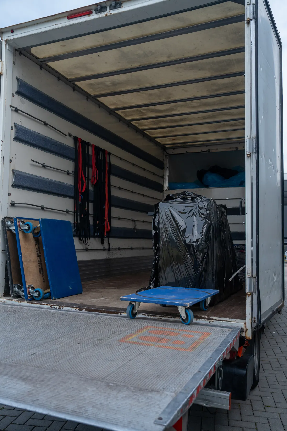 Geopende verhuiswagen met laadklep, blauwe rolplateaus, spanbanden aan de wand en een in zwarte folie gewikkeld stuk