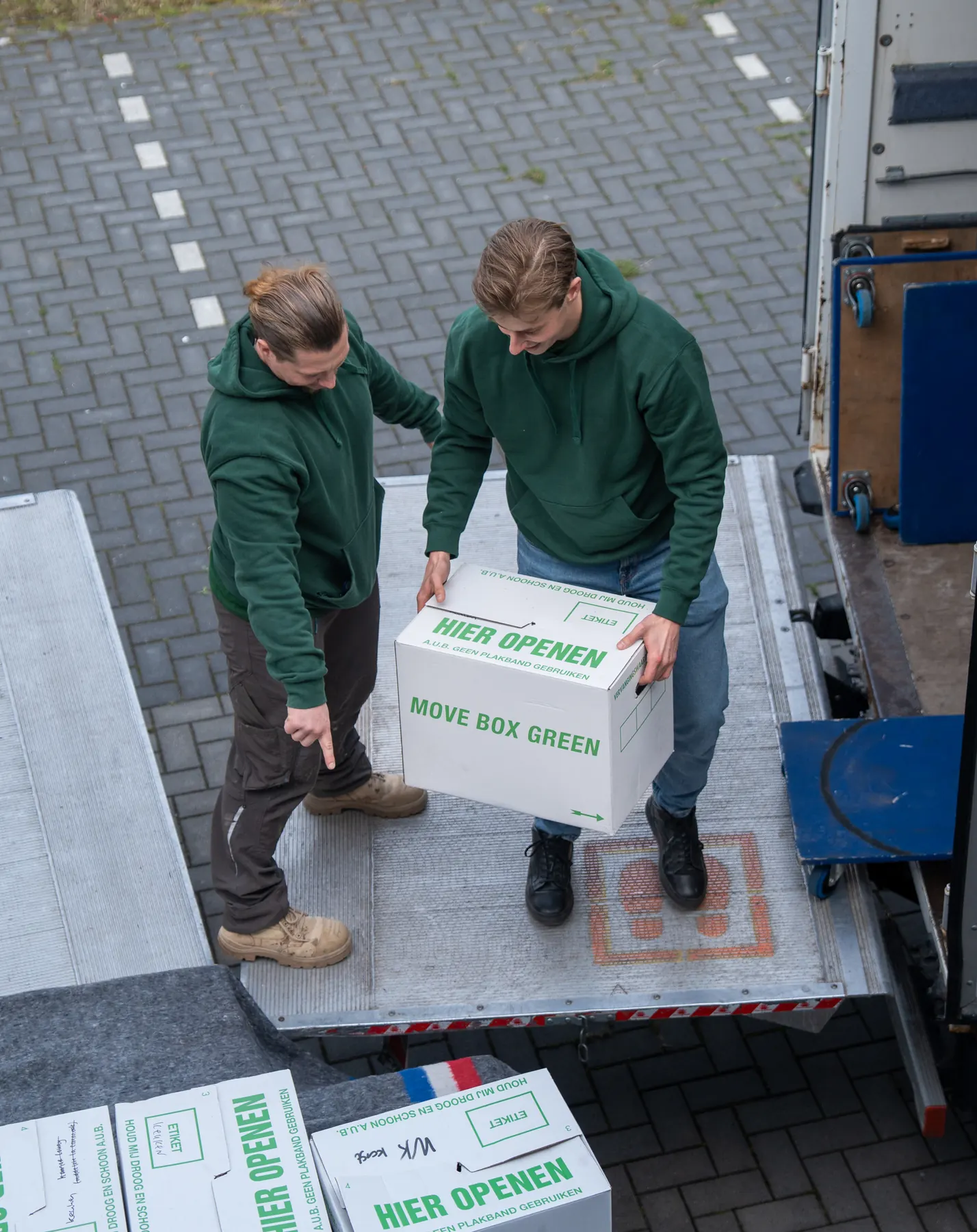Twee verhuizers van VerhuisMeesters met een verhuisdoos op de laadklep, van bovenaf gezien, met dozen met een handgeschreven etiket op de voorgrond