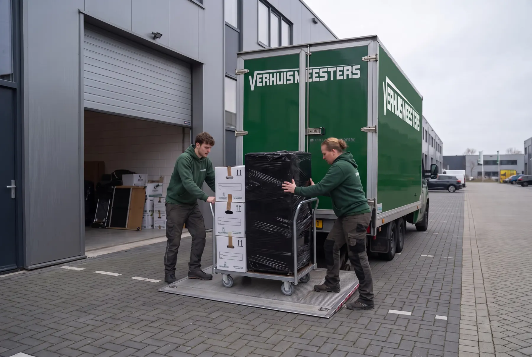 Groene VerhuisMeesters-vrachtwagen bij een bedrijfsunit, twee verhuizers met een ingepakte kast op de laadklep
