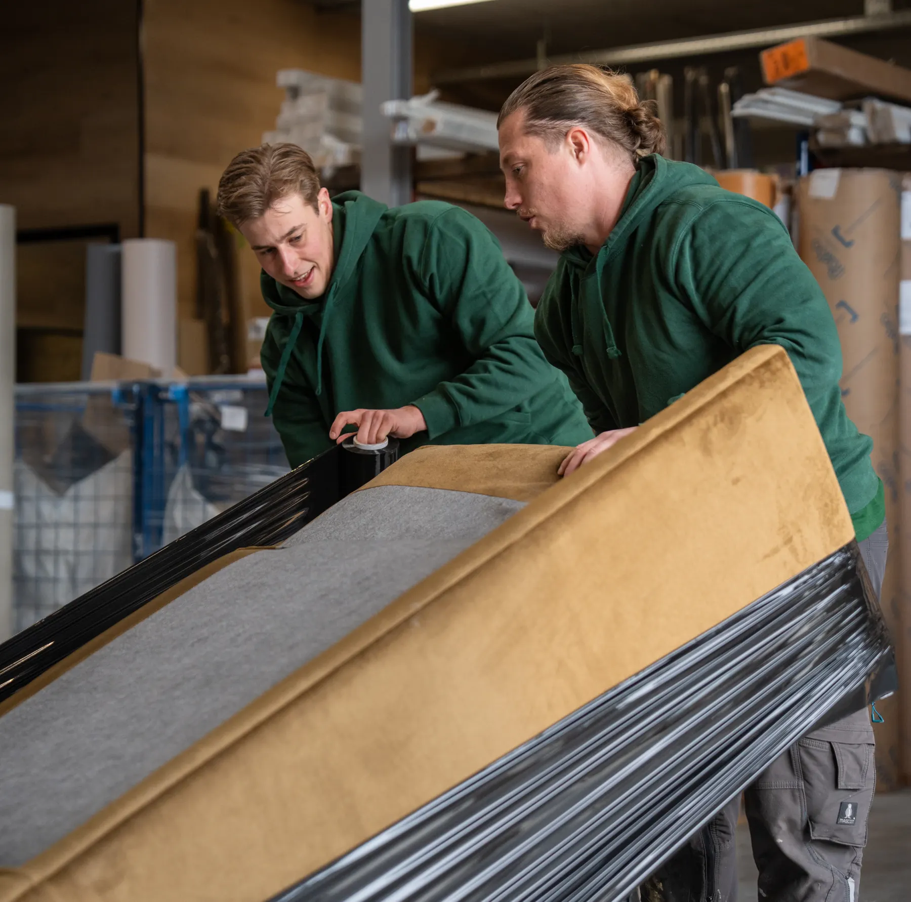 Twee verhuizers in groene werkkleding wikkelen een bank met okergele fluwelen bekleding in zwarte folie, in een loods met stellingen
