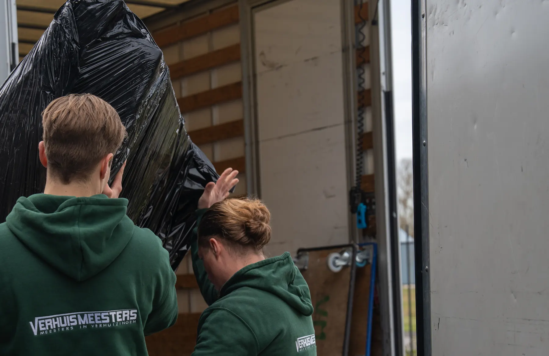 Twee verhuizers van VerhuisMeesters tillen een in zwarte folie verpakt stuk inventaris de laadruimte van een verhuiswagen in