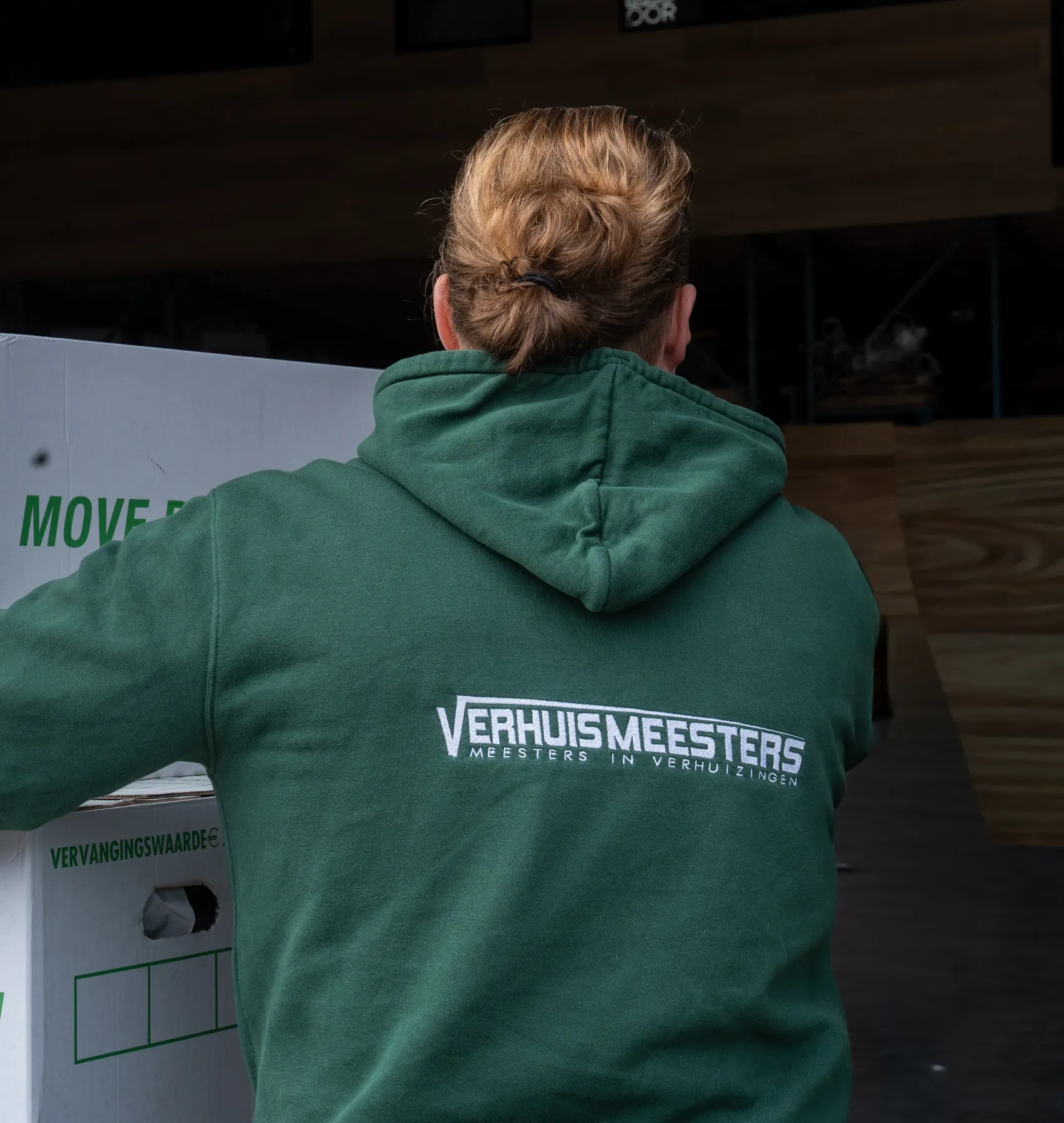Verhuizer in een groene VerhuisMeesters-hoodie, van achteren gezien, bij gestapelde Move Box Green-verhuisboxen