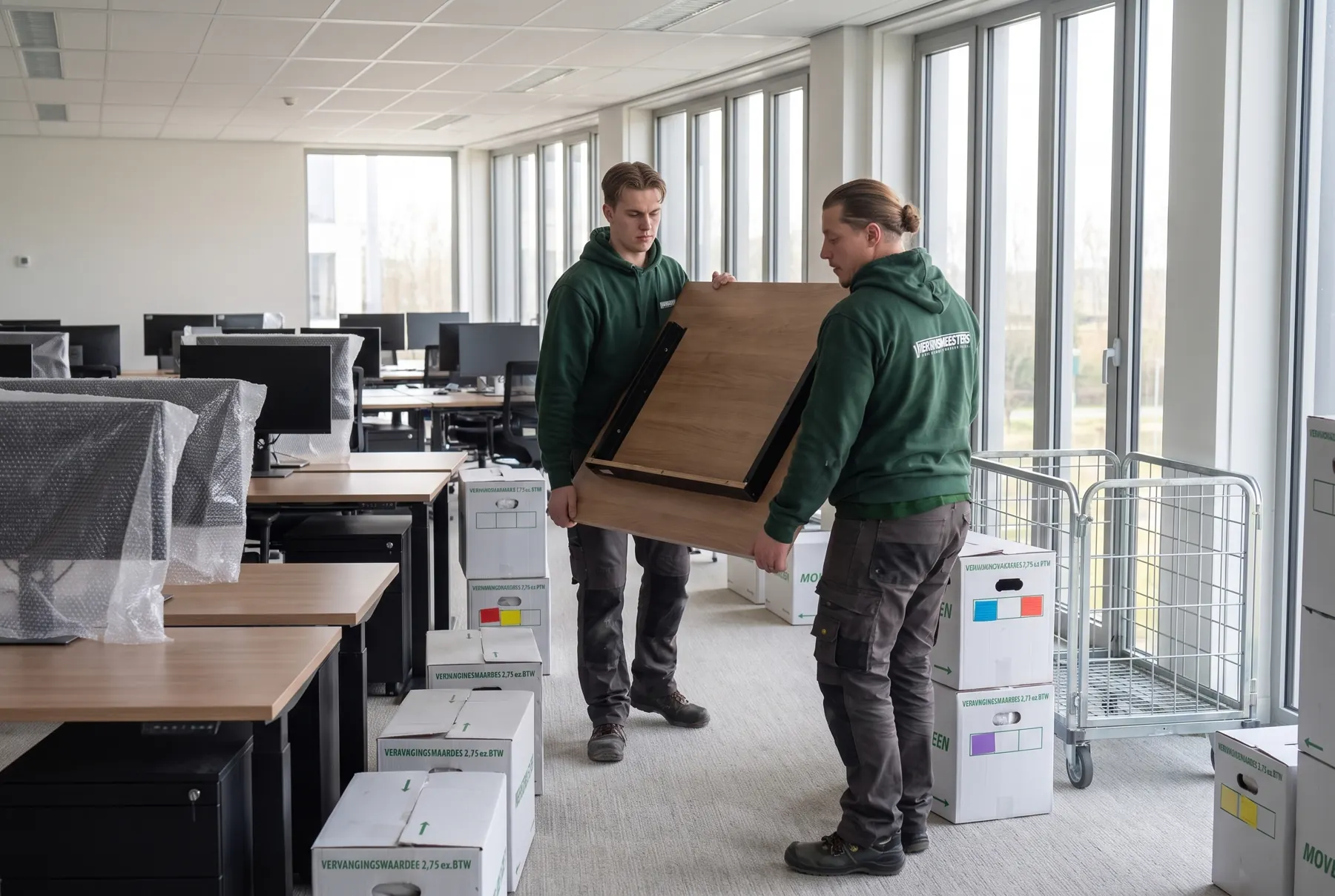 Twee verhuizers van VerhuisMeesters dragen een bureaublad door een licht kantoor vol ingepakte werkplekken