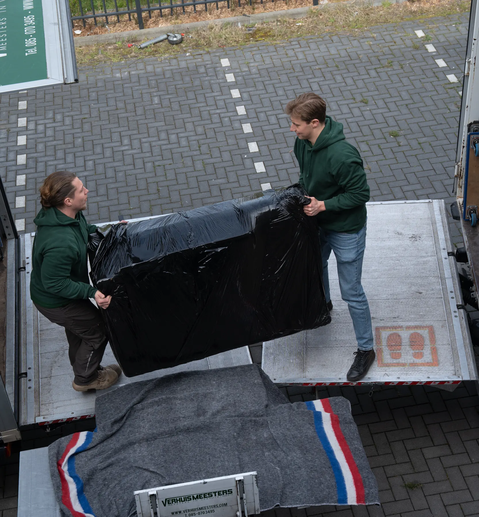 Van bovenaf: twee verhuizers tillen een groot stuk in zwarte folie over de laadklep, boven een verhuisdeken op de verhuislift