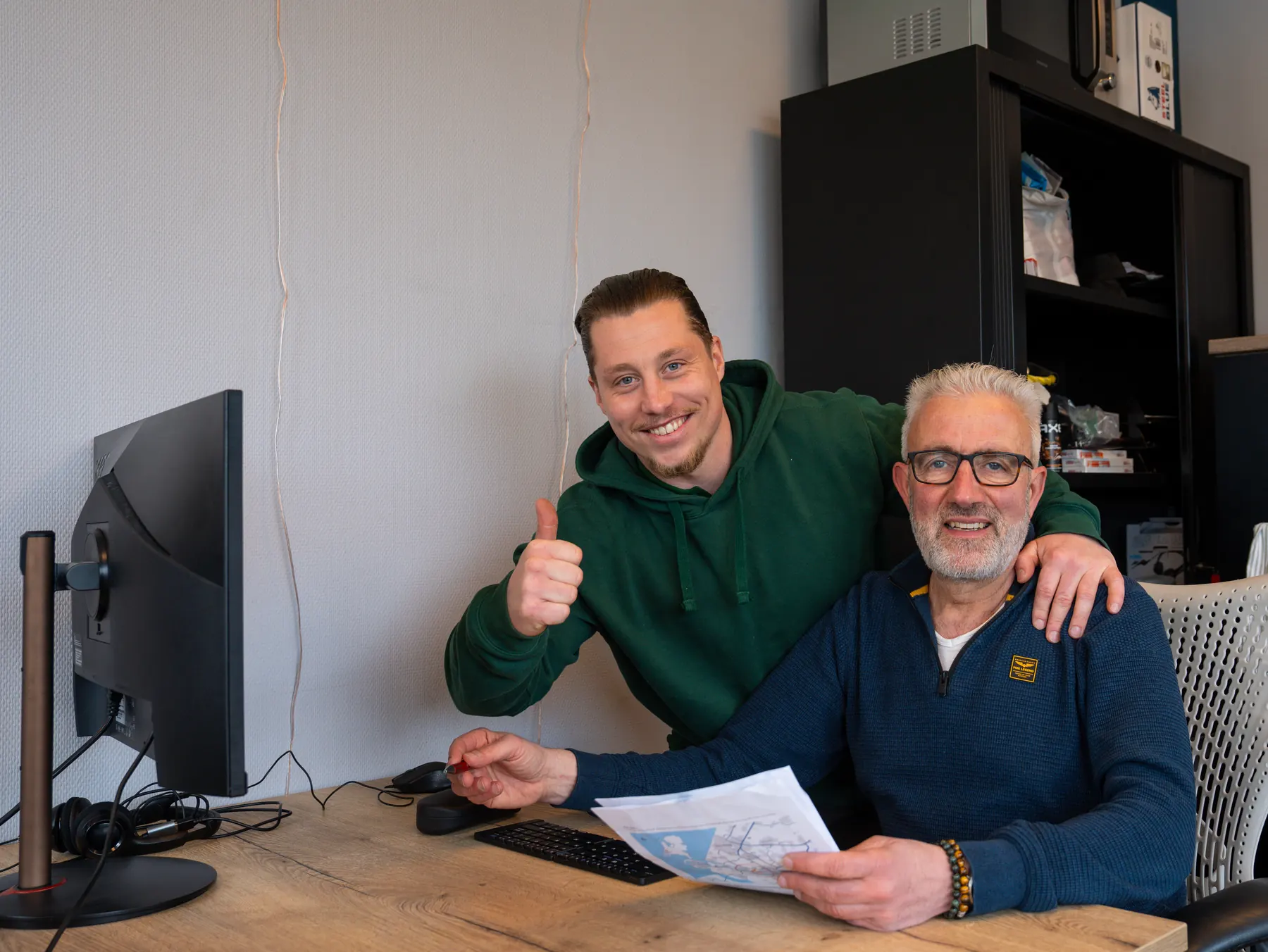 Stevie, eigenaar van VerhuisMeesters, steekt zijn duim op naast een man die aan een bureau een plan op papier vasthoudt
